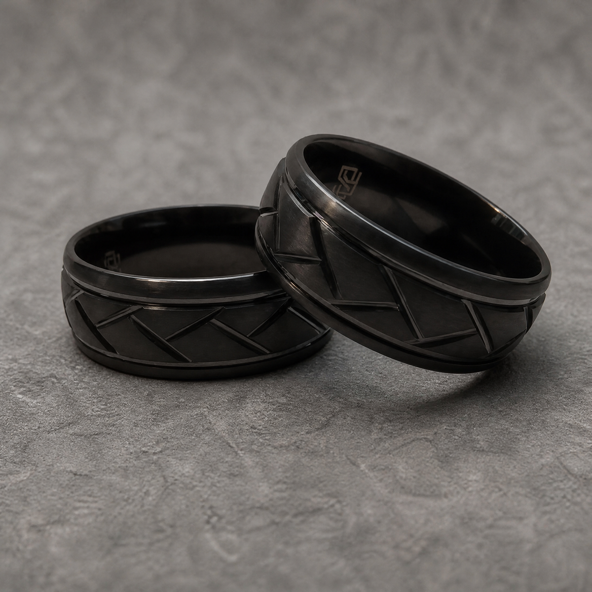 Deux bagues Ravenstone Shadow Ridge en tungstène noir à motif entrelacé géométrique, posées l'une contre l'autre, gravure "RS" visible à l'intérieur, sur fond gris texturé.