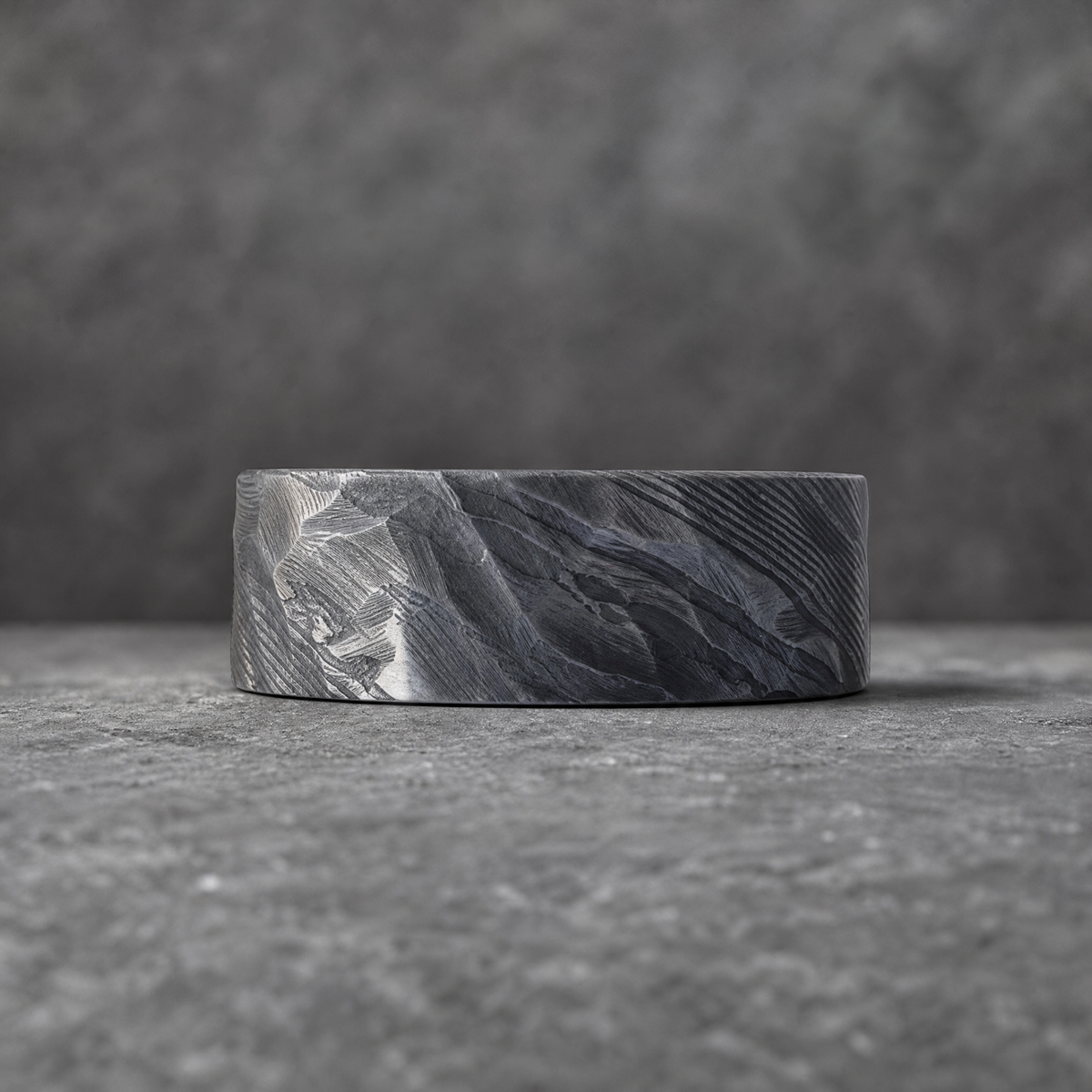 Bague Ravenstone Valhalla en acier de Damas au motif ondulé, couchée sur le côté, vue rapprochée sur fond de pierre grise floutée.