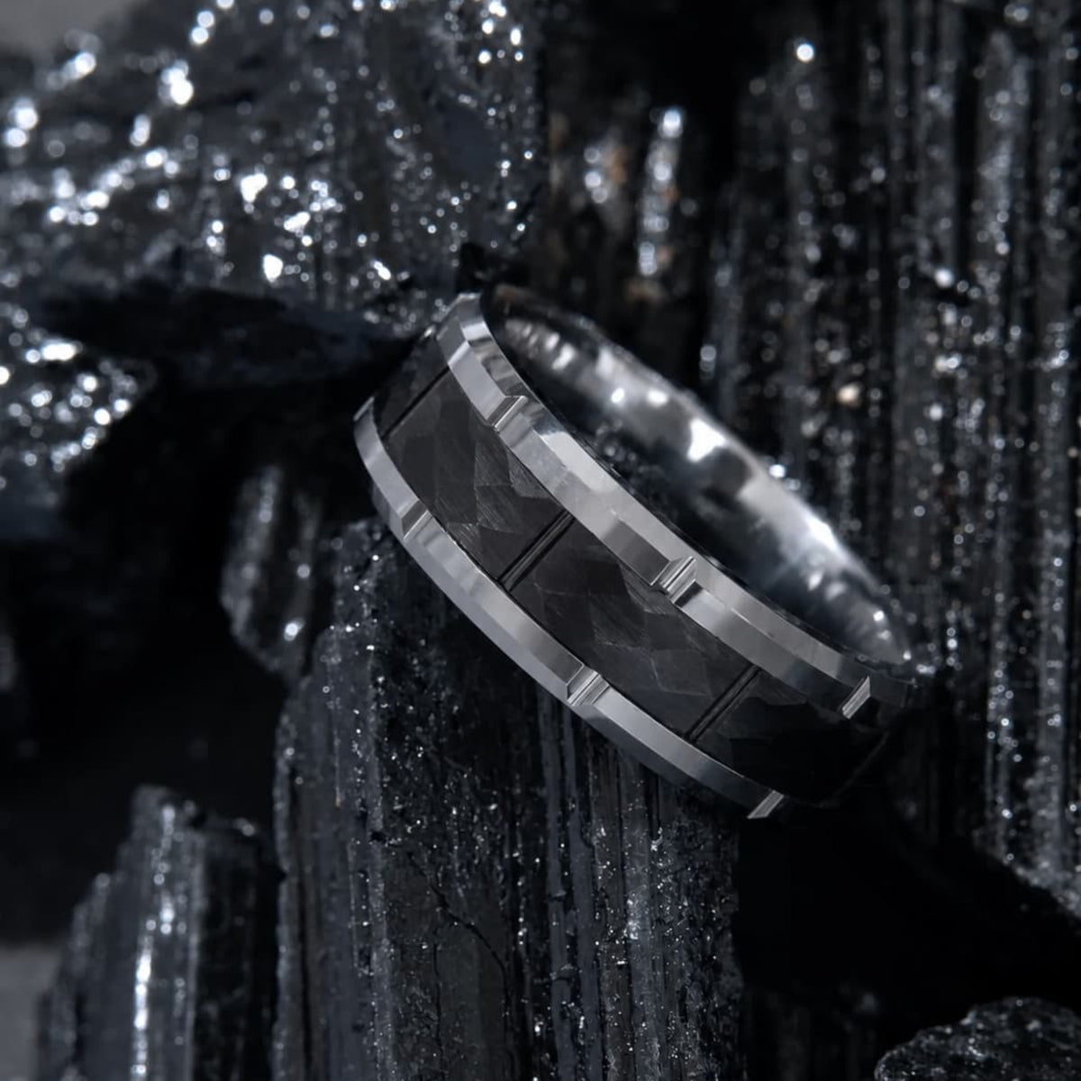 Bague Ravenstone en tungstène facettée noire avec bord argenté, posée sur roches cristallines noires.
