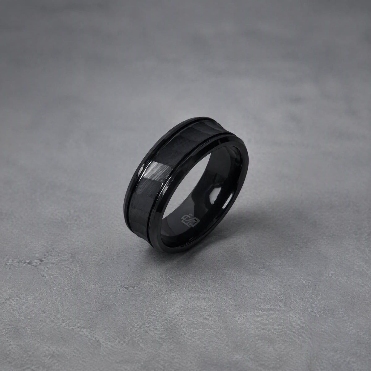 Bague Ravenstone en tungstène facetté noir, posée sur fond gris texturé.