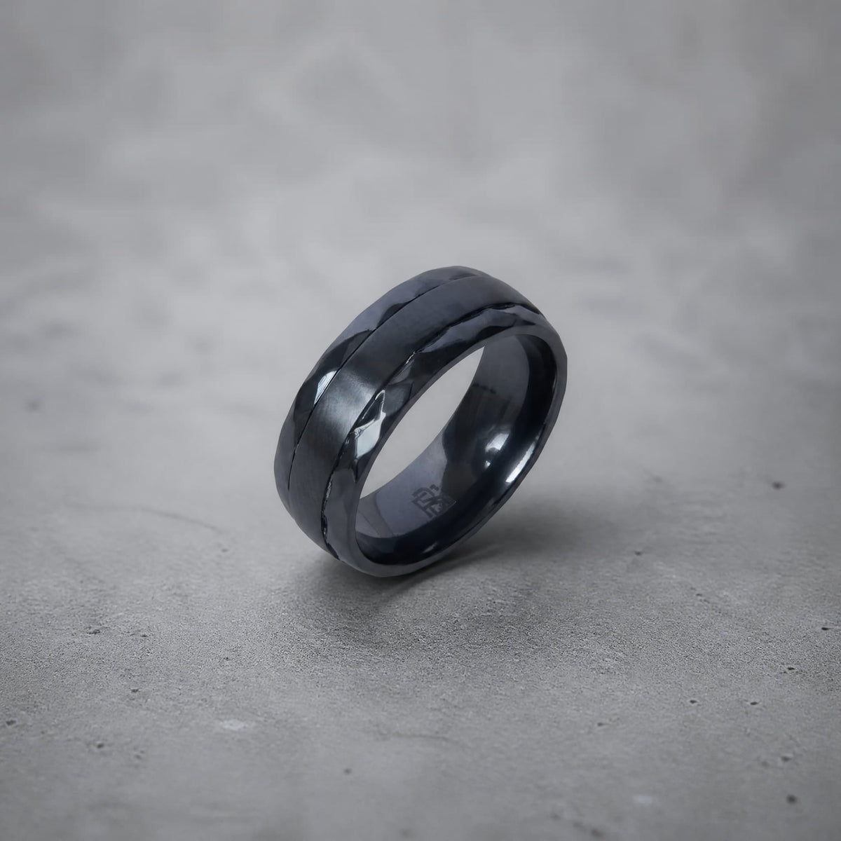 Bague Ravenstone en zirconium noir avec bande lisse centrale et bords facettés, posée sur fond gris lisse.