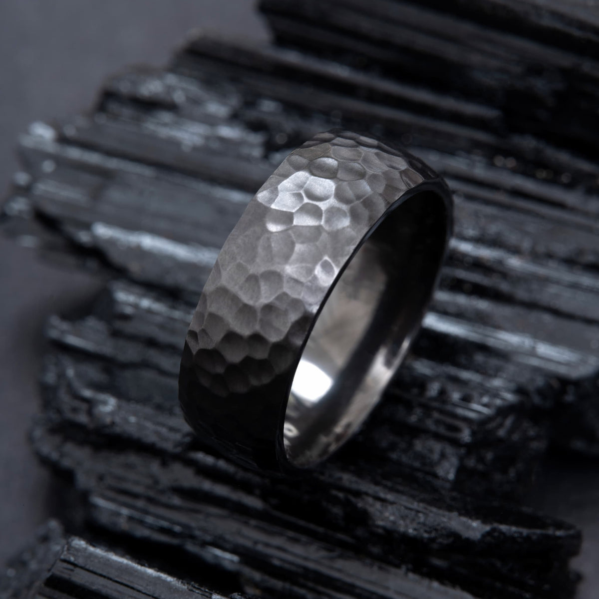 Bague Ravenstone en zirconium noir martelé, posée sur des roches sombres.
