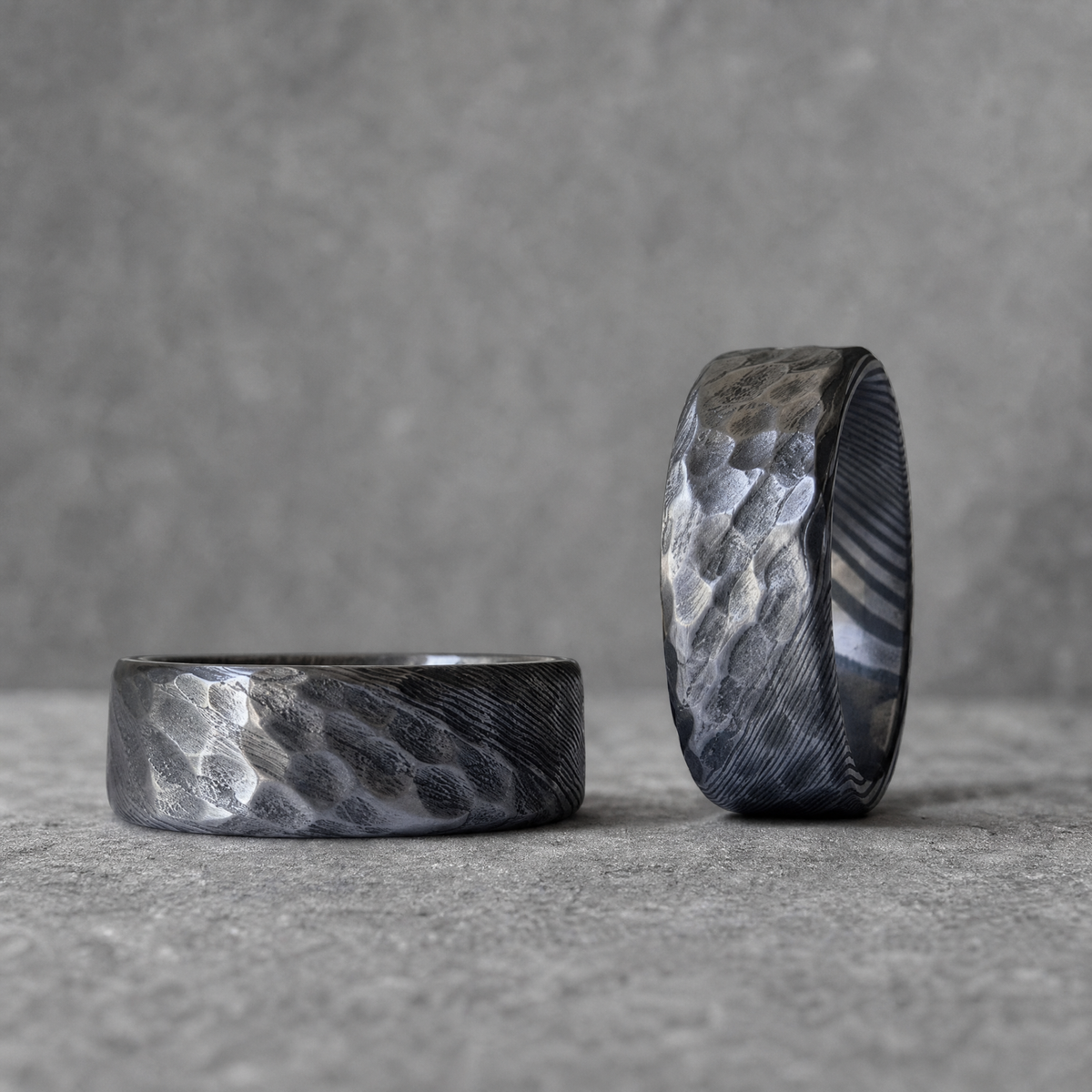 Deux bagues Ravenstone Legacy en acier de Damas à double texture martelée et striée, une couchée au premier plan et une debout, gros plan sur fond gris.