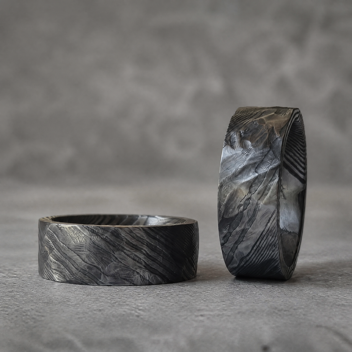 Deux bagues Ravenstone Valhalla en acier de Damas au motif ondulé, une debout à l'arrière et une couchée au premier plan, sur fond gris.