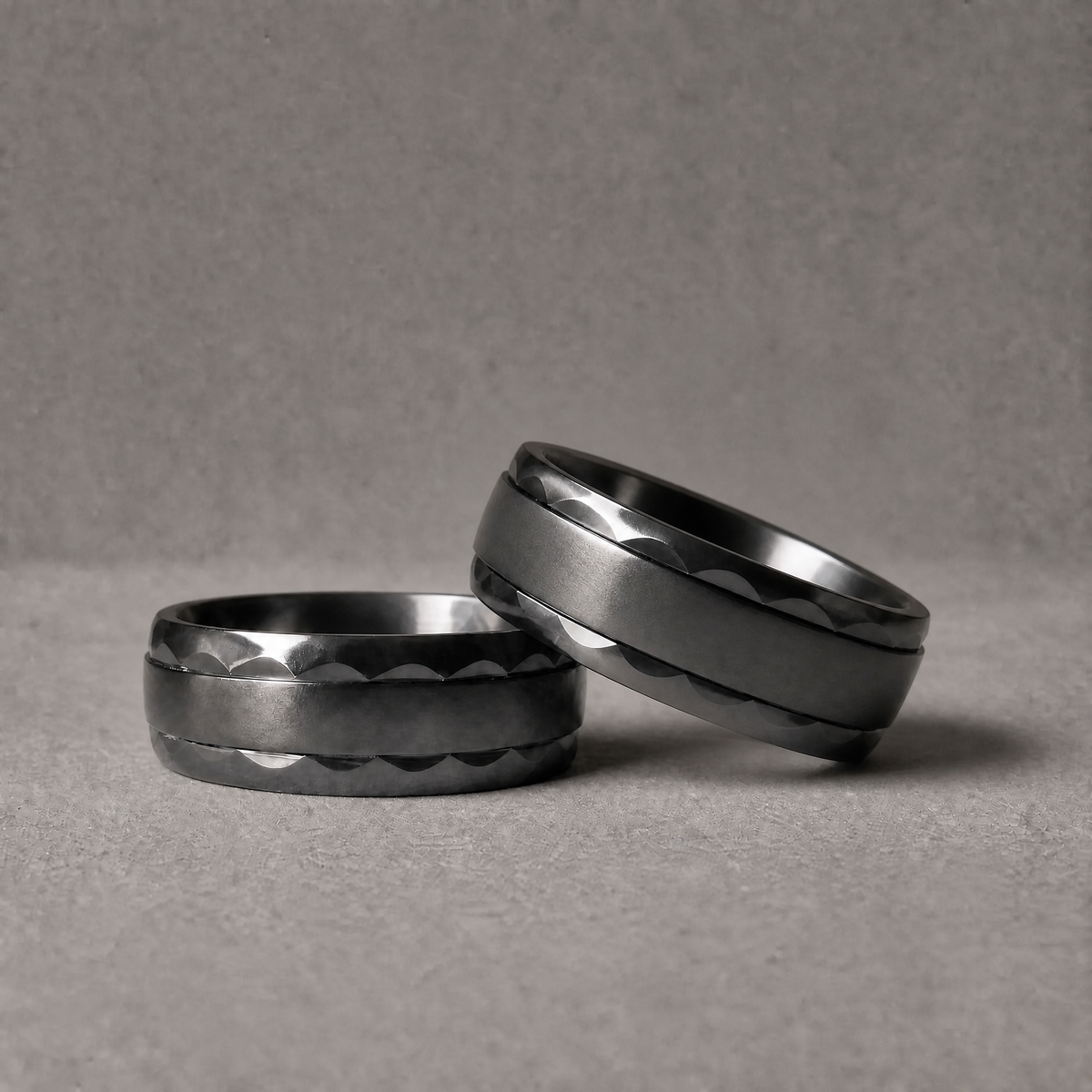Deux bagues Ravenstone Phantom en zirconium noir à motif facetté et bords biseautés polis, posées inclinées l'une contre l'autre, sur fond gris.