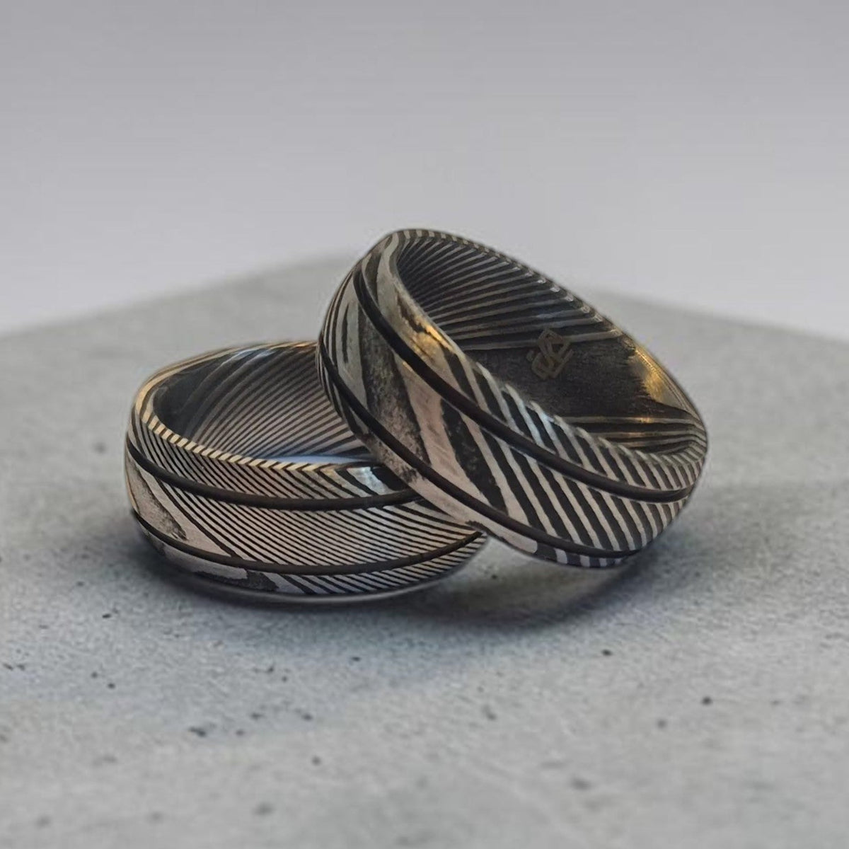 Deux bagues en damas noir aux motifs striés contrastés, posées sur une surface grise texturée. 