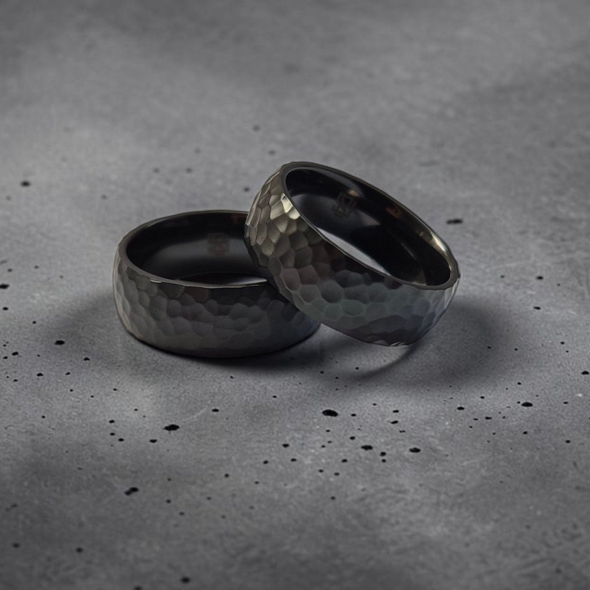 Deux bagues en zirconium noir à texture sculptée, posées sur une surface grise brute. Les reliefs irréguliers du métal créent une présence dense et minérale, révélant une esthétique sombre, contemporaine et résolument architecturale.