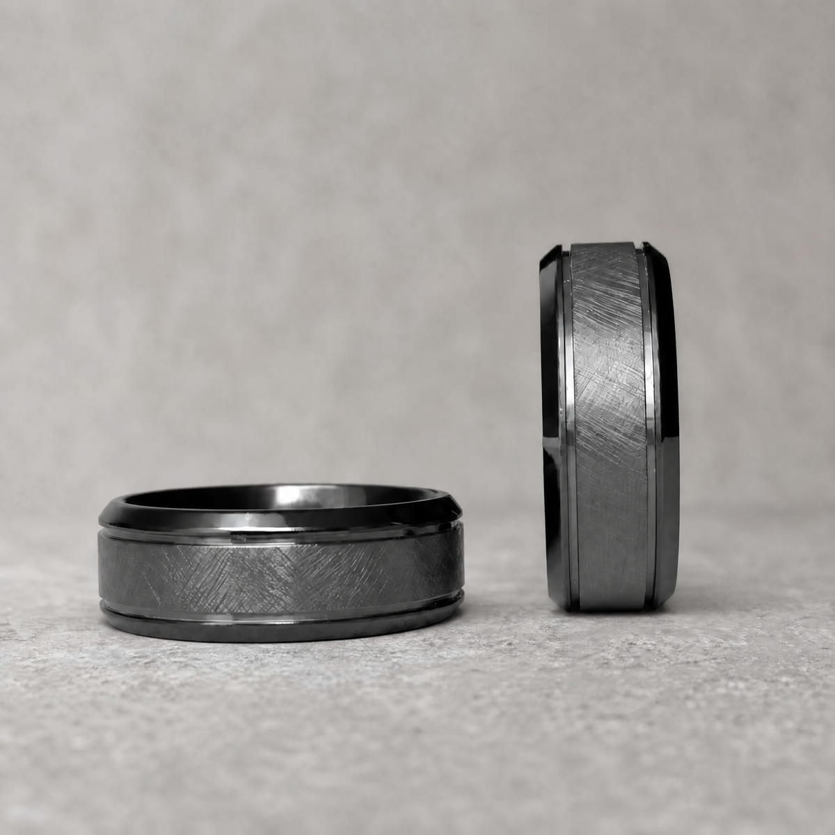 Duo de bagues Ravenstone Nightstone en zirconium noir à texture brossée et bords biseautés polis, une bague posée à plat, l'autre debout, sur surface grise texturée.