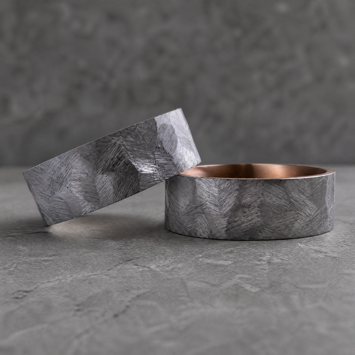 Duo de bagues Ravenstone Aurum en tantale gris-bleu à texture gravée chevrons, une bague appuyée contre l'autre laissant voir l'intérieur en or rose poli, fond gris texturé.