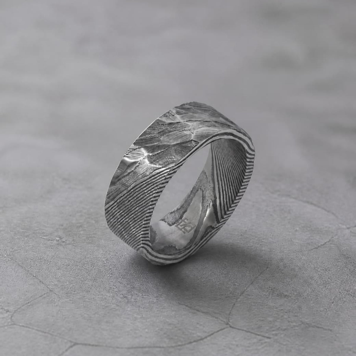 Bague masculine en acier damas gris anthracite, avec texture martelée et motifs métalliques striés, photographiée sur un décor minéral gris éditorial.
