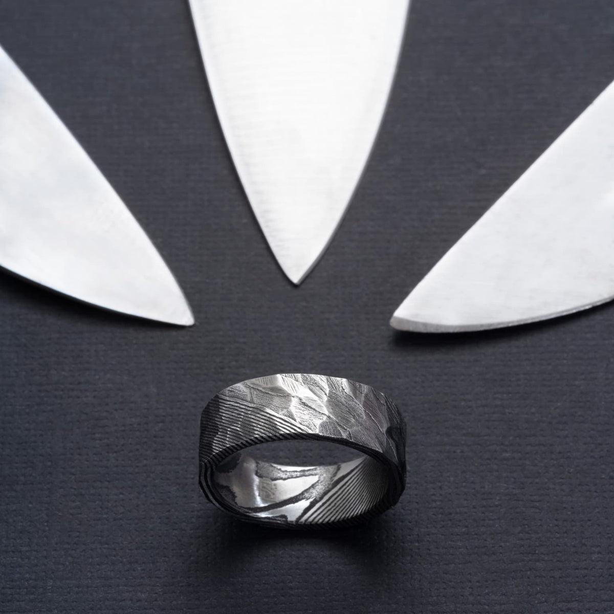 Bague homme en acier damas gris anthracite, ornée d’un motif martelé et de stries diagonales, présentée sur une surface noire entourée de lames.