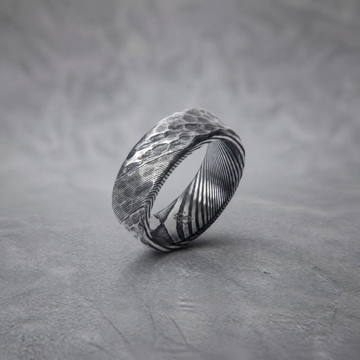 Bague Ravenstone en acier de damas martelé, posée sur un fond gris texturé.