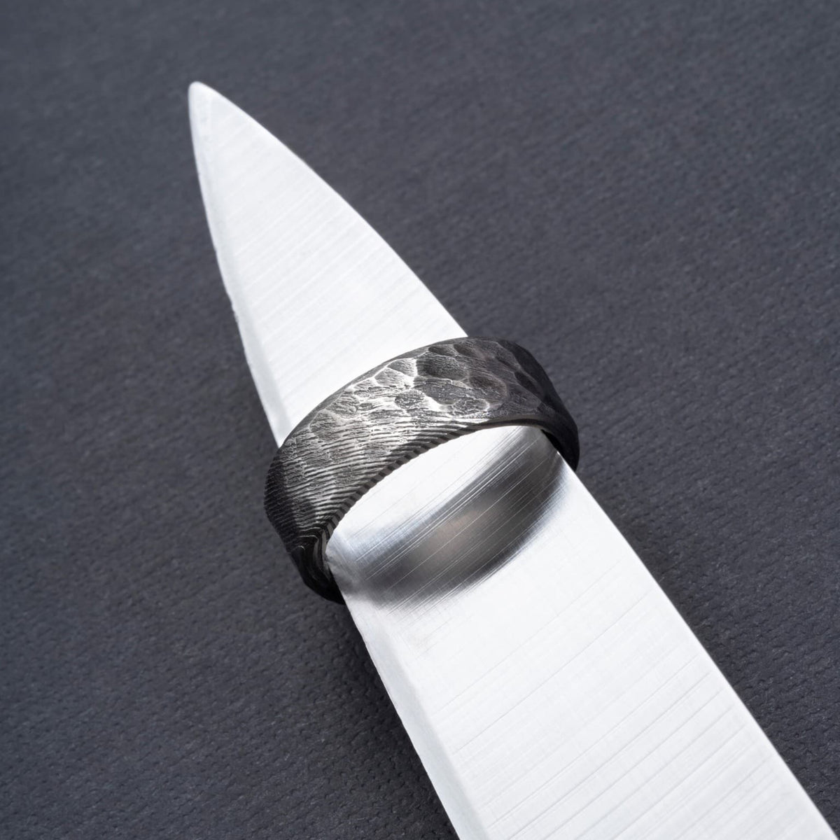 Bague Ravenstone en acier de damas martelé, posée sur une lame métallique sur fond sombre.