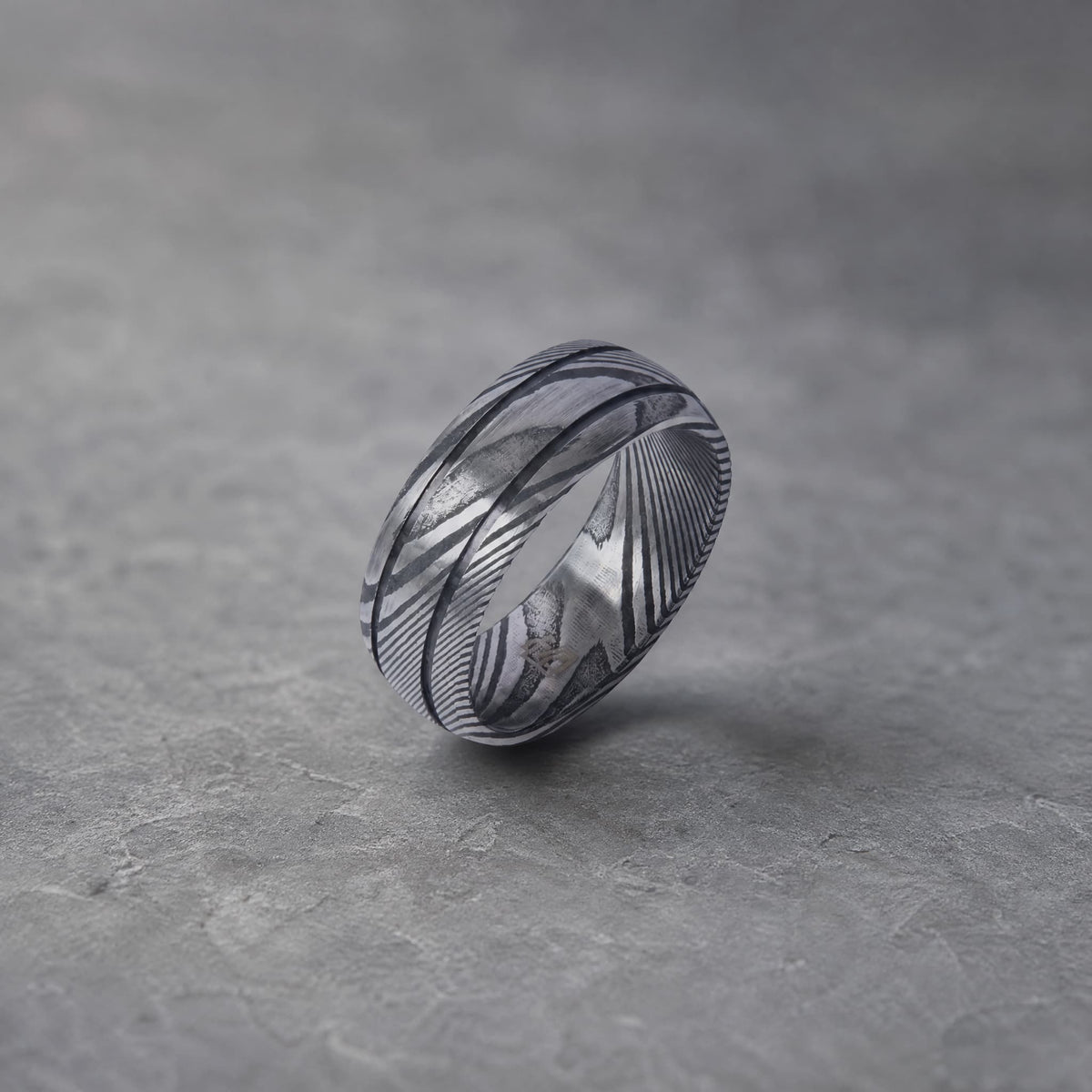 Bague Ravenstone en acier damas à rainures, posée sur un fond gris texturé.