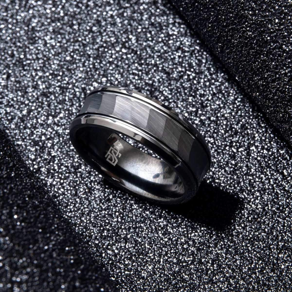 Bague Ravenstone en tungstène noir brossé, faccetée, posée sur un fond noir scintillant.