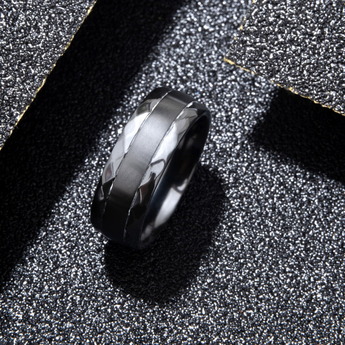 Bague Ravenstone en zirconium noir avec bande lisse centrale et bords facettés, posée sur un fond noir scintillant.