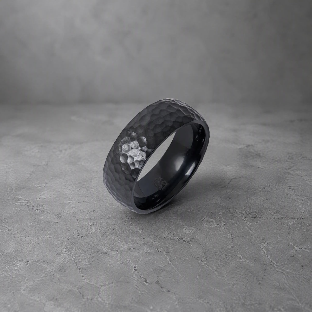 Bague Ravenstone en zirconium noir martelé, posée sur un fond gris effet béton.